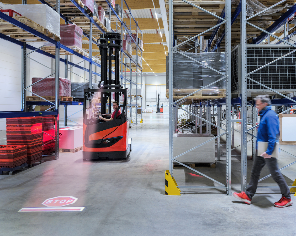 A man walking n a warehouse down one aisle and a Lined high reach forklift driving down another aisle